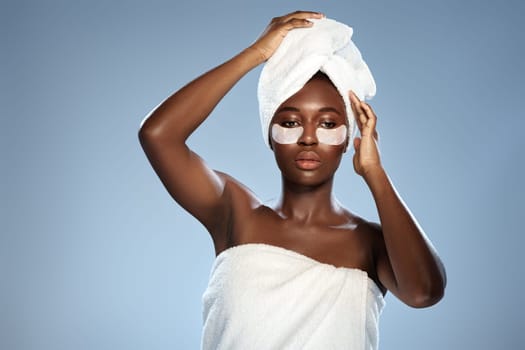 A woman with glowing skin is applying an eye treatment while dressed in a towel. Her hair is wrapped in a towel, creating a relaxing atmosphere in a stylish space.