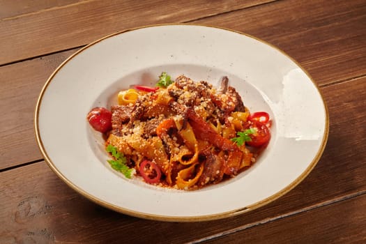 Pappardelle with meat and vegetables, decorated with grated cheese and herbs, served in a white plate on a wooden table
