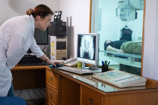 A radiologist in a white lab coat sitting at a workstation while analyzing an X-ray image of a patients hip in a medical imaging room.