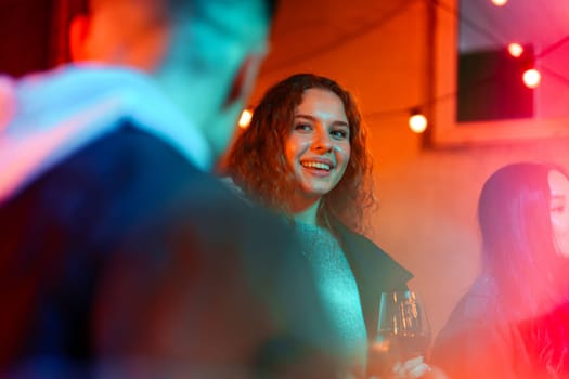 A woman with curly hair smiles brightly as she engages in conversation with friends at an outdoor gathering adorned with colorful lights, creating a fun atmosphere during the evening.