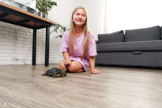 A joyful girl wearing a purple shirt interacts with her pet turtle on the wooden floor of a bright living room. Sunlight filters through the window, creating a warm atmosphere.