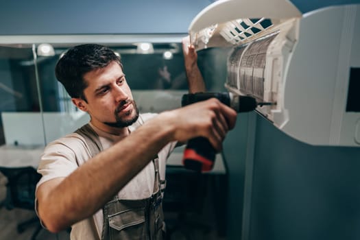 A technician is focused on installing an air conditioning unit inside a contemporary office space. He is using a power drill to secure the unit in place, ensuring optimal functionality.