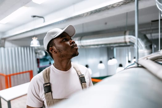 A young man wearing a cap and overalls carefully examines machinery in a contemporary industrial setting, focused on ensuring proper functionality.