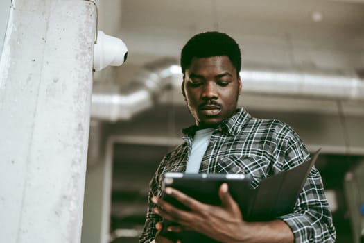 A young man dressed in a plaid shirt examines documents on a tablet in a spacious, industrial-style office with visible piping and modern decor.