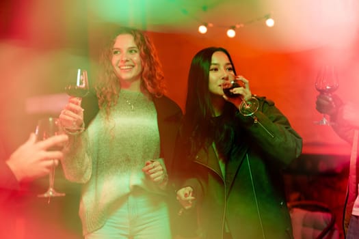 Two friends raise their glasses while laughing and enjoying a fun evening together at a cozy indoor gathering filled with colorful lights and good energy.