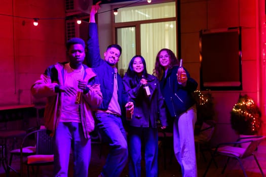Four friends enjoy a lively gathering at night, surrounded by colorful lighting and decorations. They hold drinks, celebrating and posing joyfully together.