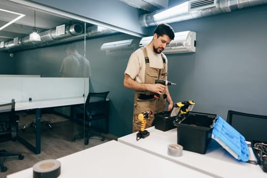 A skilled worker wearing overalls is carefully sorting tools in a workspace. The modern office features metal ductwork and clean, minimalist design, indicating ongoing repairs.
