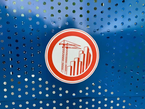 Circular red sign featuring construction cranes and skyscrapers, mounted on blue perforated metal sheet, represents restricted access and emphasizes ongoing urban development activity