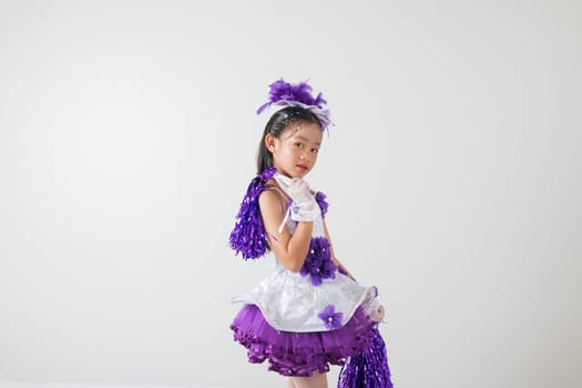 Little cheerleader girl in purple uniform with pom-poms, smiling joyfully. White studio background highlights her enthusiasm and team spirit. cheerleader kid