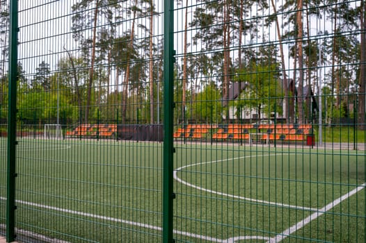 football field with white lines on the grass. With orange stands. Tall trees around. Surrounded by a green net, a fence around the football field in the form of an iron net with large cells