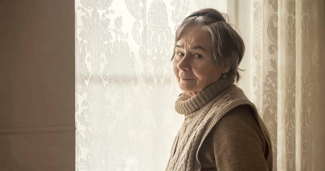 An elderly gray-haired woman in a casual knitted sweater looks out the window of her house, an old grandmother is waiting for guests, stands against the background of a window with lace curtains.