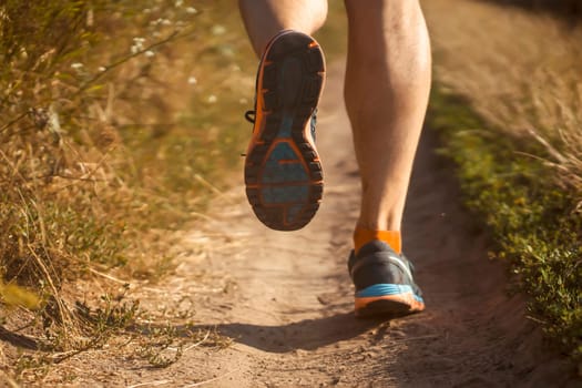 Athlete runner legs running on a close-up of a trail across a field on a shoe. Fitness and warm-up, young man with sun effect in the background.