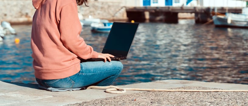 A girl is typing on a laptop keyboard, sitting and working while traveling against the backdrop of a seascape, a bay with a sea on a sunny day, a woman is screening information online on her device.