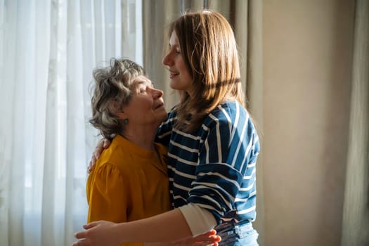 Granddaughter visits and hugs her beloved grandmother at the weekend, a happy daughter joyfully spends time together with her elderly mother at home indoors stand near the window.