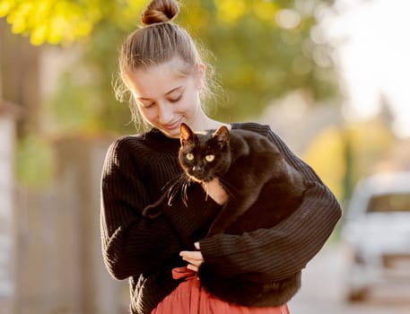 Pretty girl in red skirt hugging black cat outdoors at street with autumn leaves. Beautiful model teenager sitting with feline animal at park