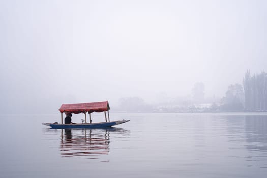 Srinagar, Kashmir - 3rd Jan 2025: Man sitting in a red roofed Shikara wooden boat on Dal lake famous place with floating market cafes etc on a foggy day
