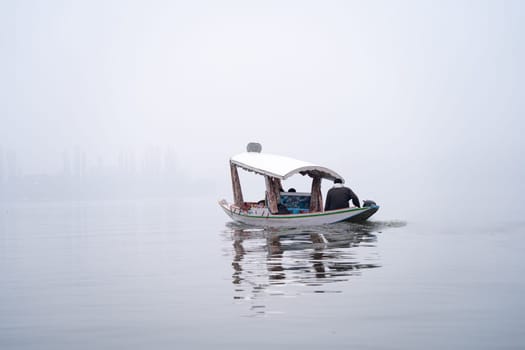 Wooden Shikara boat being rowed on the serene blue waters of Dal lake in the middle of winter fog during the months of Chilai Kalan in Srinagar Kashmir India