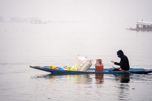 Boatman rowing a wooden canoe with gas cylinders and food items on dal lake showing the water transport based local economy in Srinagar Kashmir