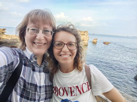 Two friends capture a joyful selfie by the seaside, enjoying their travel adventure in a picturesque coastal location under a clear sky.
