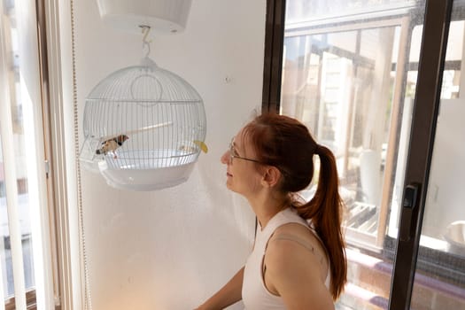 Redhead woman admiring her pet parrot in a white cage hanging from the ceiling