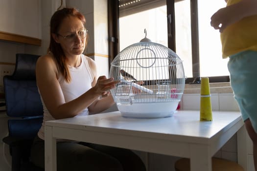 Woman changing the water in a bird cage while a child watches in a home setting