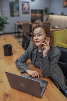 Woman on phone with laptop at table. She wears a grey blazer in a bright, modern, spacious office. Potted plant and art are visible.