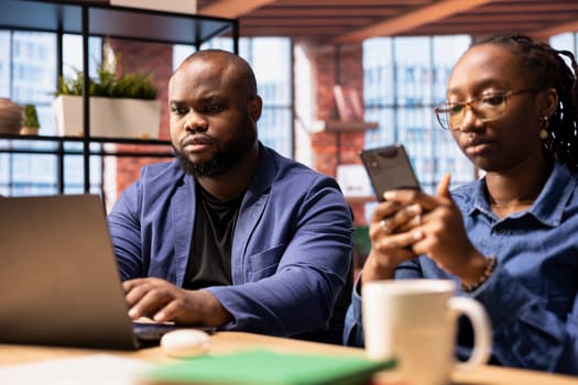 African American couple in a modern flat collaborate on a consulting project, using technology to enhance their productivity. Man works on laptop while his partner answers emails on phone.