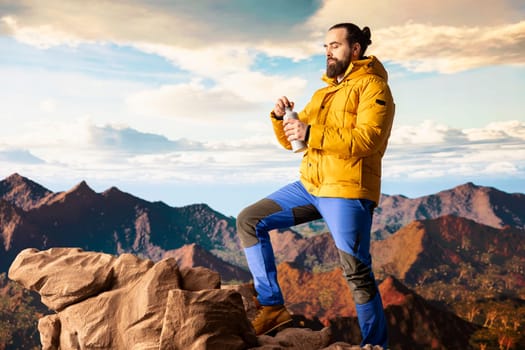 Young man explorer climbing the mountain range and hydrating with his water bottle, drinking refreshment on top of the rocky mountain. Male hiker refreshing after the trekking activity.