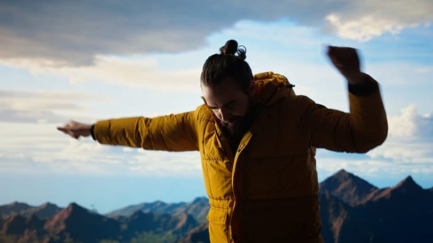 Pleased hiker celebrating his personal achievement on mountain top, showing determination for beautiful landscape and sightseeing. Adventure seeker doing a dance with satisfaction. Camera A.