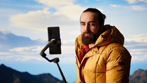 Traveler filming his hiking experience in the wilderness documenting his adventure on social media using his smartphone. Recording the beautiful scenic mountain view behind him for content. Camera B.