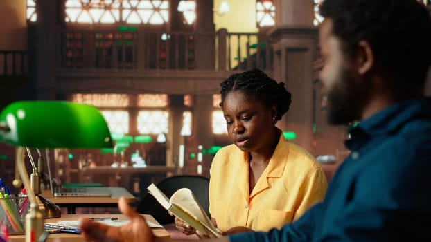 African american scholars reading course notes and comparing information at the library, studying for a test together using academic database for research. Educational materials. Camera B.