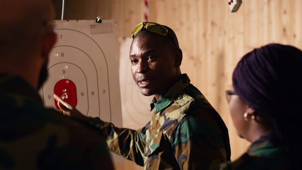 Army commander teaching recruits in shooting gallery how to shoot paper targets, explaining pointing system. Military officer instructing soldiers on how to hit bullseye, reading rules, camera B