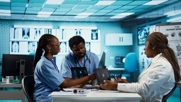 African american general practitioner leading a healthcare team in a cabinet, analyzing MRI imaging results and providing accurate diagnosis and effective treatment. Medical facility. Camera B.