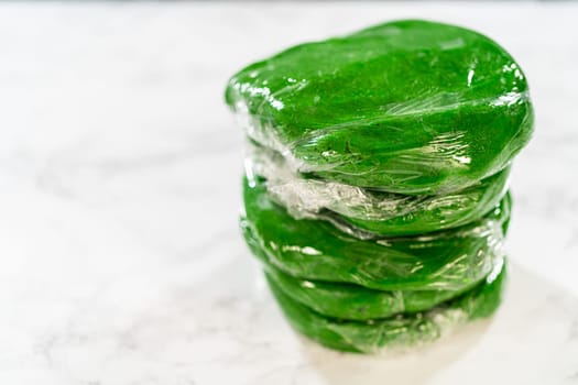 Stacked portions of green dough, each wrapped in plastic wrap, prepared for chilling in the fridge for Vibrant Green Gingerbread Cookies for Festive Baking.