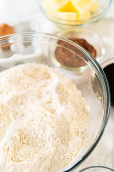 A large glass mixing bowl filled with sifted flour, placed on a marble surface along with other ingredients like spices, butter, and molasses for the Chocolate Gingerbread Cookie Dough.