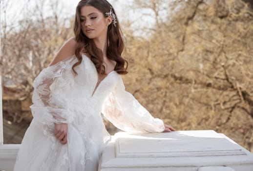 Young brunette bride in traditional long dress posing on white hotel balcony in early spring. white dress with long sleeves, corset, open shoulders. Portrait of the bride, wedding makeup and hairstyle