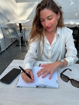 A woman with long hair is focused on writing in her notebook at a cafe terrace during daylight, enjoying a quiet moment.