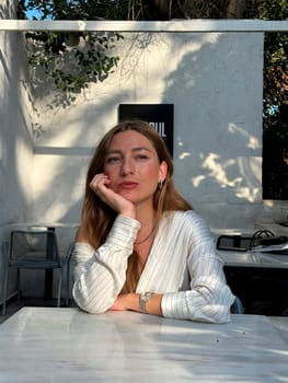 A young woman relaxes at a white table in a cozy outdoor cafe, basking in the sunlight while deep in thought.