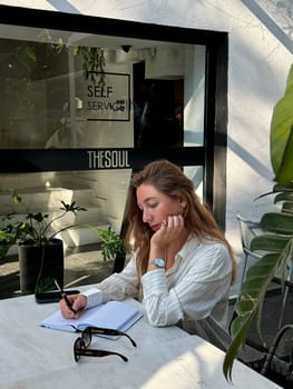 A woman sits at a table in a cafe surrounded by greenery, focused on writing in her notebook, enjoying the peaceful atmosphere.