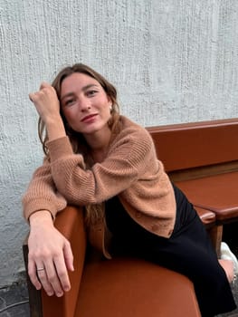 A young woman leans comfortably on a brown bench, showcasing a relaxed pose, in a bright urban environment.
