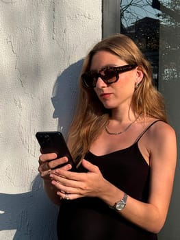 Young woman wearing sunglasses interacts with her smartphone, enjoying the afternoon sun by a stylish wall.