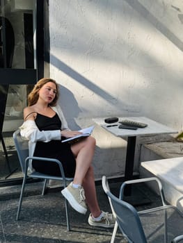 A woman sits comfortably at a cafe table, writing in a notebook, enjoying the warm afternoon sun. She wears a casual outfit and appears focused.