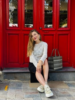 A young woman relaxes on steps near a bright red door, dressed in a stylish outfit, enjoying the warm day.