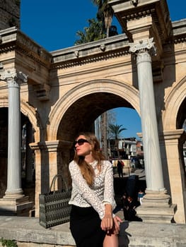 A young woman relaxes under grand arches in a historic location, surrounded by palm trees and lively atmosphere during daytime.