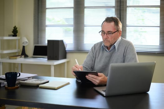 Focused elderly male working on tablet beside laptop in modern home office.
