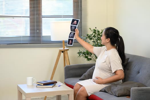 Happy pregnant woman admiring sonogram photos and gently touching her belly.