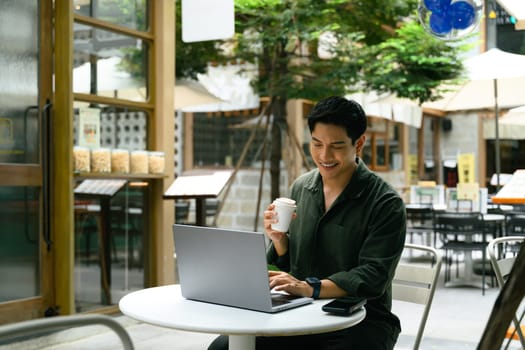 Smiling man working on a laptop at an outdoor cafe and enjoying a cup of coffee.