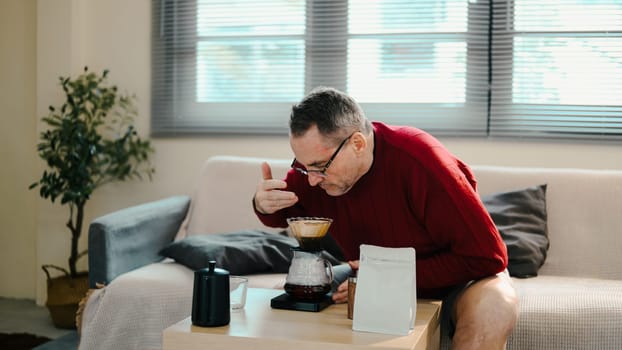 Older man in red sweater smelling coffee brewed with pour-over set on wooden table.