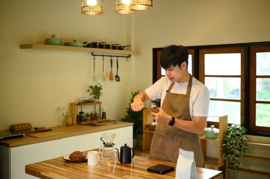 Young man in apron grinding fresh coffee beans in a home kitchen.