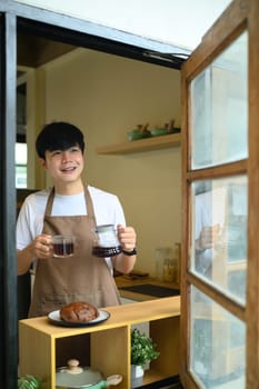 Young man holding a fresh cup of coffee and carafe, smiling through a window.